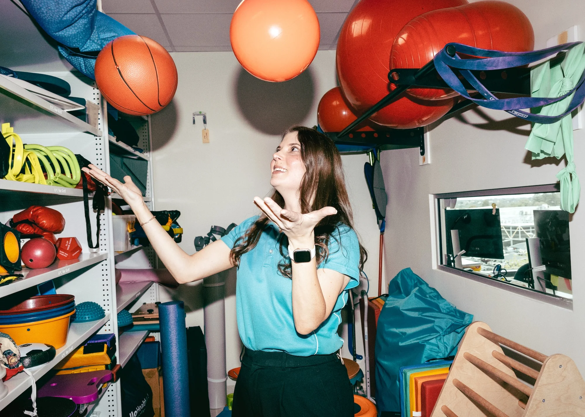 An occupational therapist in a therapy equipment room tossing a ball in the air, surrounded by shelves of exercise balls, mats and mobility aids